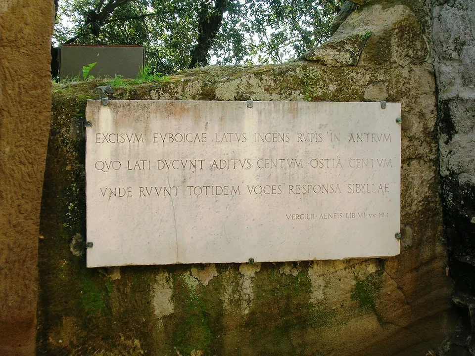 A white marble plaque fixed to a mossy tufa wall, carved with three lines of Latin in capitals and a smaller line naming Virgil's Aeneid, book VI.
