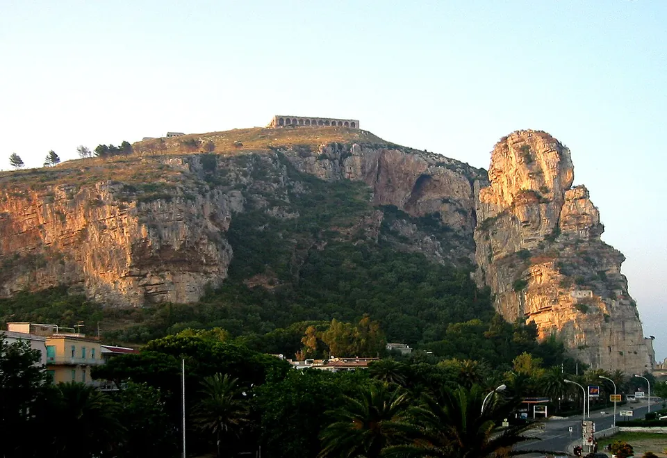 A long ridge of pale limestone cliffs lit by low sun, with a separate rock pinnacle at its right-hand end, a row of arches running along the top of the ridge, and trees and the edge of the modern town at its foot.