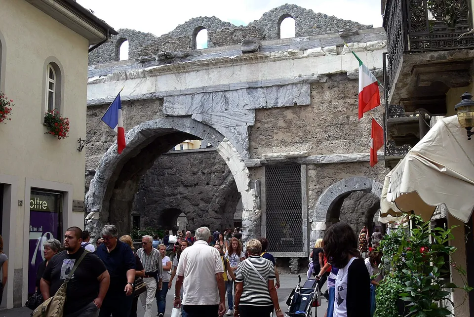 A busy street passing through a large round stone arch beside a smaller one, the masonry topped by a band of pale stone slabs and a ruined upper wall with small arched windows, flags hanging from the houses on either side.