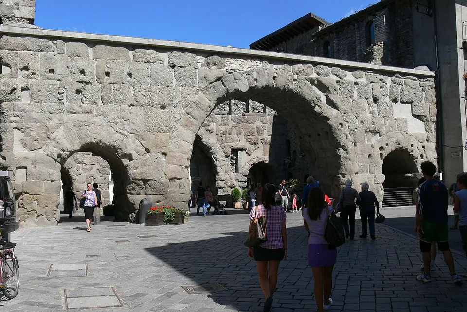 A broad wall of large rough stone blocks pierced by one big round arch between two smaller ones, with a further row of arches visible through them, and people walking across the paved square in front in bright sun.
