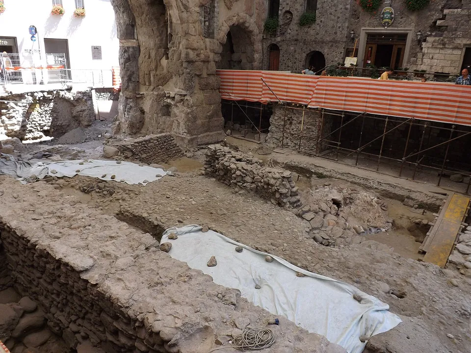 An excavation trench inside the gate, with stone wall foundations, sheeted areas and scaffolding under a striped awning against the arches and the houses beyond.