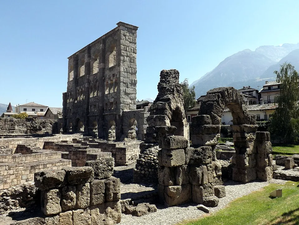 Broken stone piers and the stumps of arches stand among low walls and gravel in front of the tall façade, with houses, trees and a mountain behind.