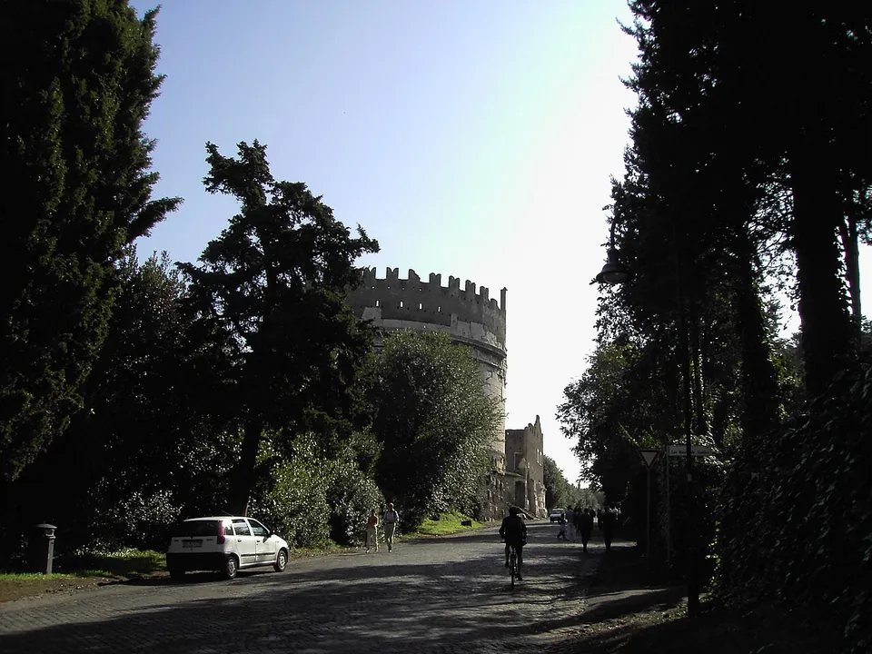 A paved road runs between tall trees towards a round crenellated tower at the roadside, with a parked car on the left and people walking and cycling on the road.