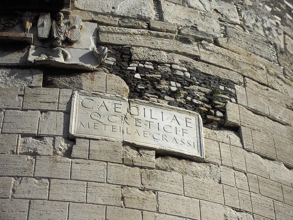 A rectangular marble plaque set into a wall of squared stone blocks, carved in three lines CAECILIAE, Q CRETICI F, METELLAE CRASSI, with a weathered relief above and to the left.