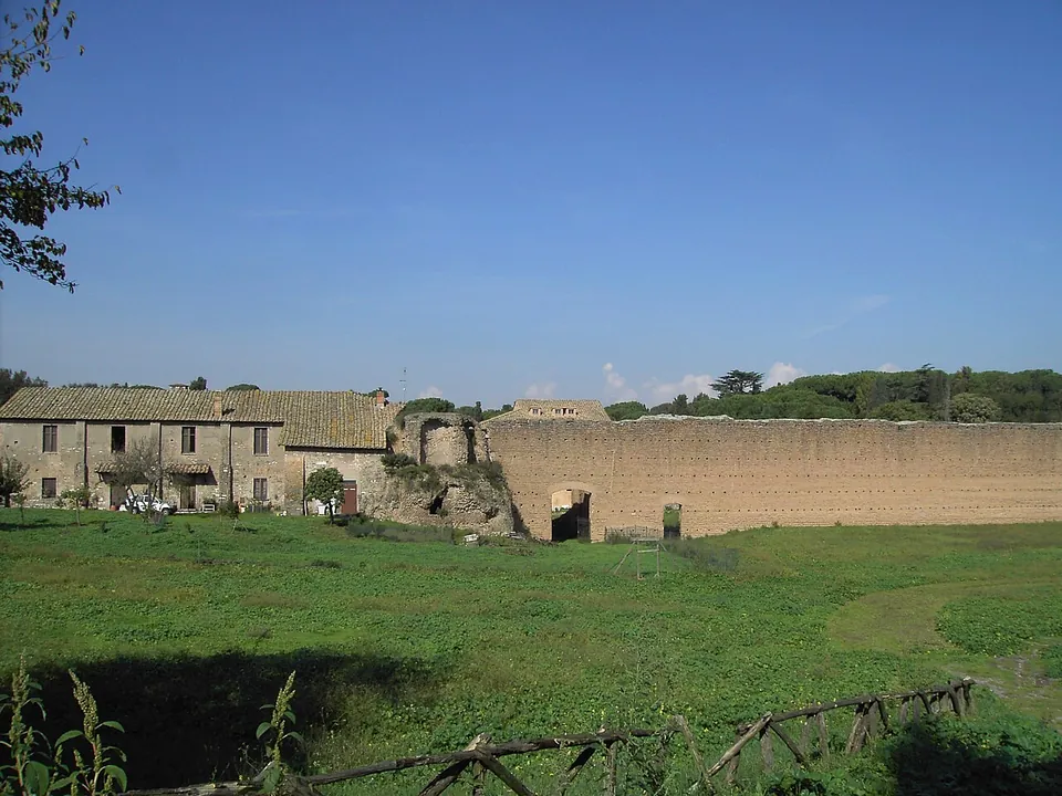 A long, low farmhouse with a tiled roof on the left, a ruined brick structure with an arch beside it, and a high wall of brick and stone running off to the right across a green field.