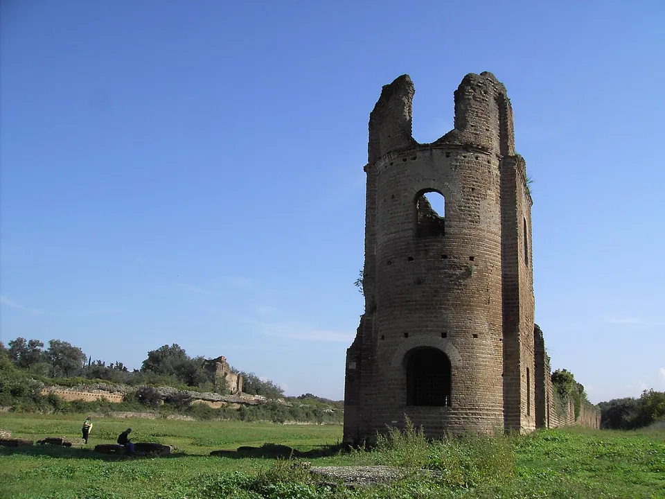 A tall ruined tower of brick, with an arched doorway at its foot and an arched window above, its top broken into two stumps; a long, low wall runs away from it across a grassy field.