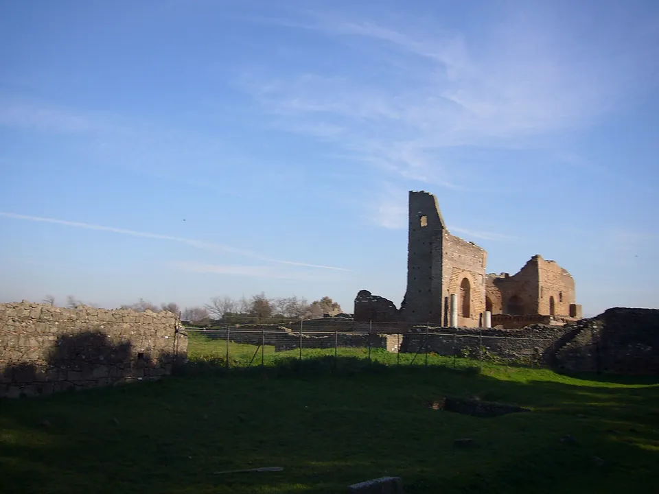 Across a grassy field and a low fence, a tall narrow brick ruin stands beside a lower brick building with arched openings, with a stone wall in the foreground on the left.