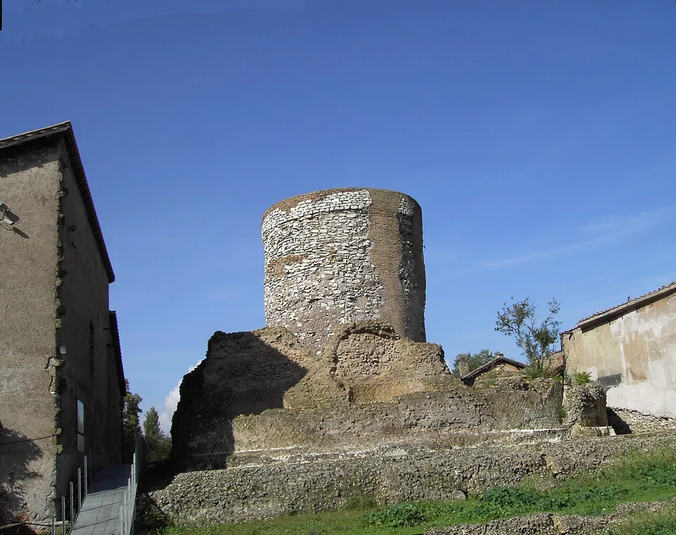 A squat cylinder of rough grey and white rubble masonry standing on top of a broad, ruined mass of older masonry, with a low grassy bank in front and the plain end walls of two farmhouses pressing in on either side, under a blue sky.