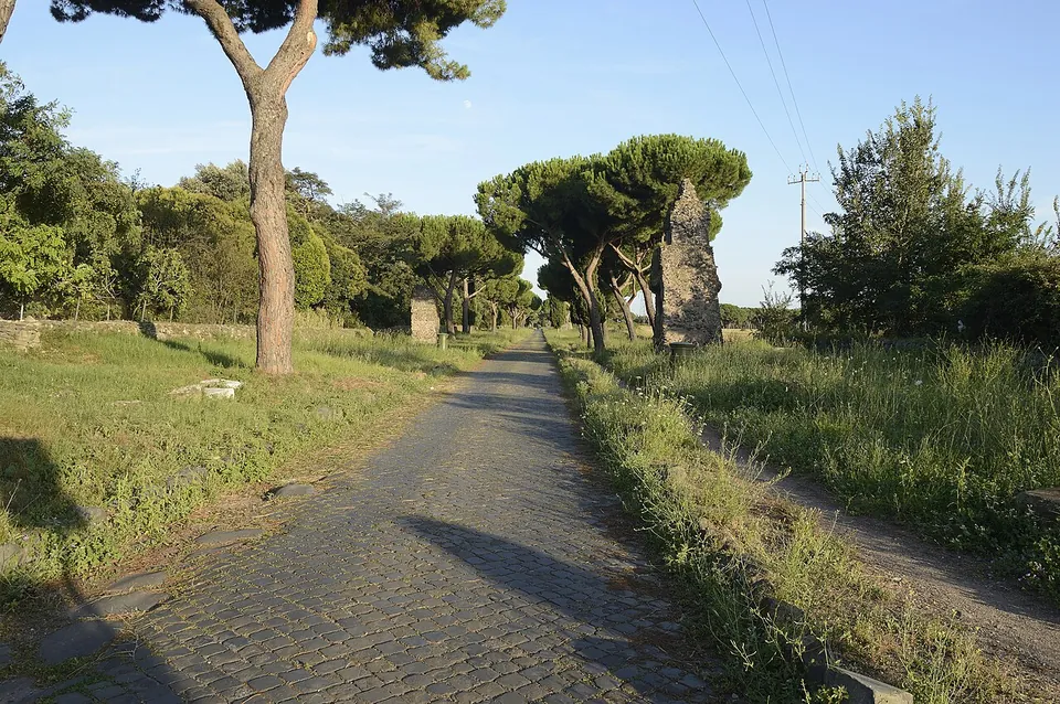 A straight road paved with large dark polygonal stones between grass verges and umbrella pines, with a tall fragment of ruined masonry at the roadside.