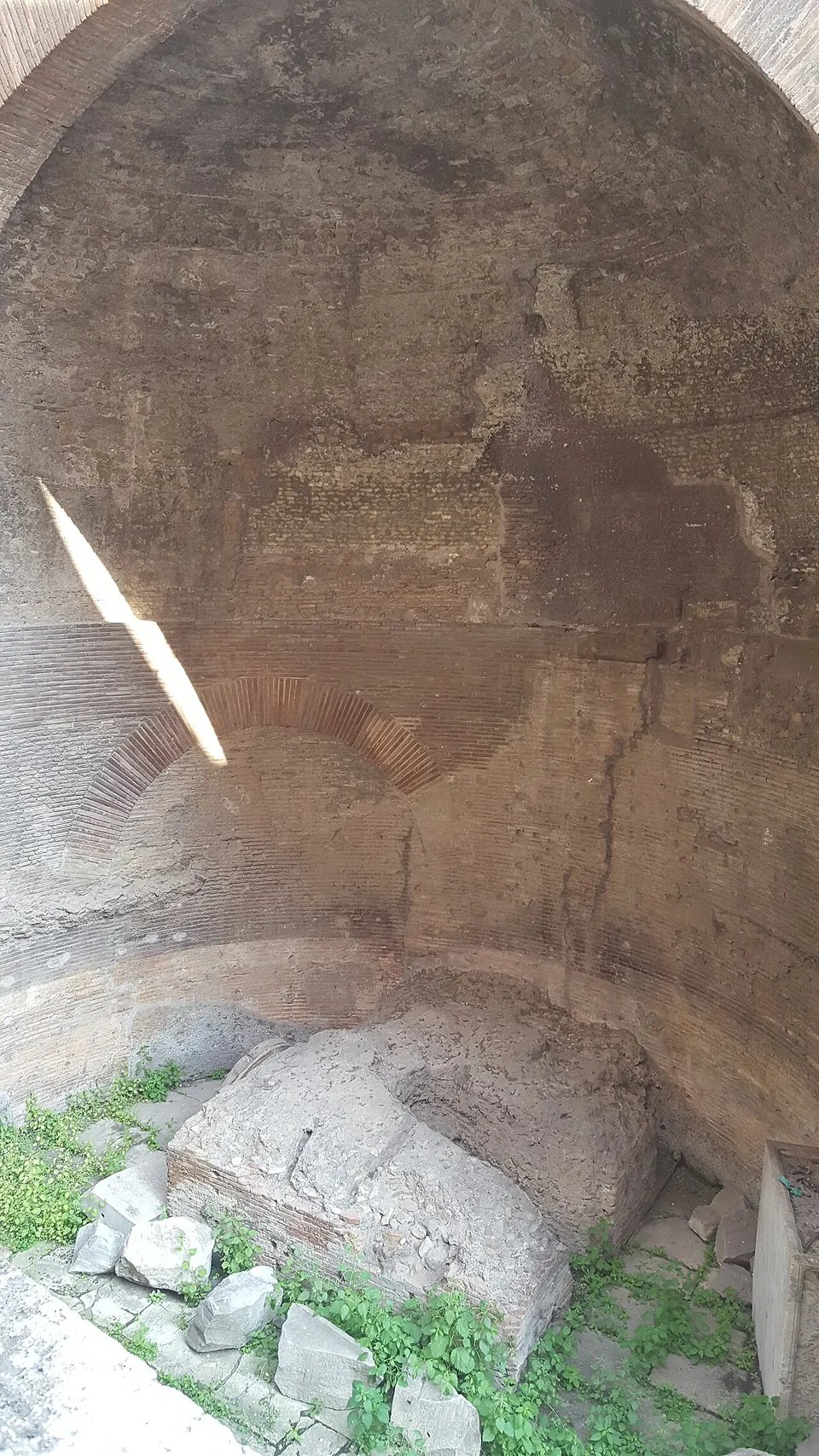 A curved brick wall rising into a half-dome, a brick arch set low in the wall and a block of brick and rubble at its foot among weeds and loose stones.