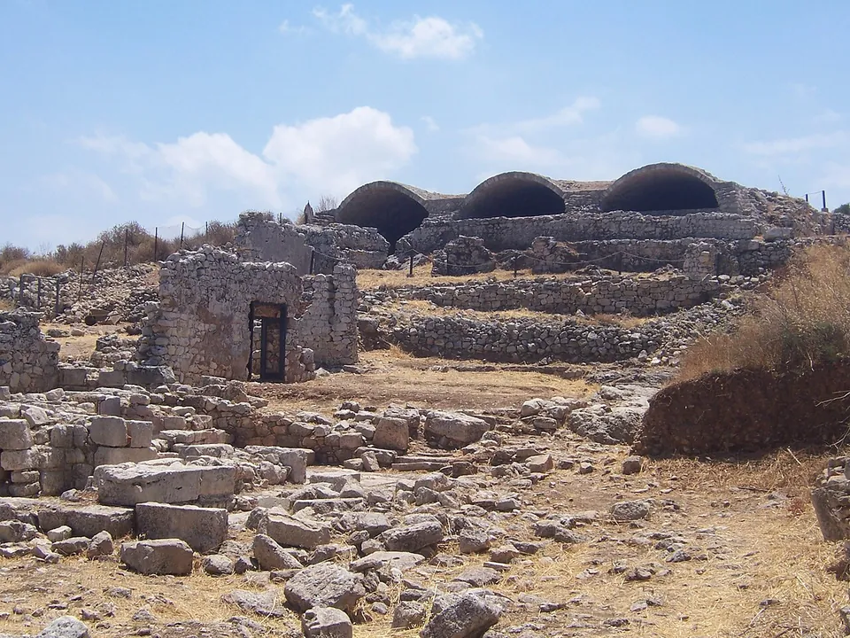 A field of tumbled stone blocks and low walls leading to a small roofless stone building with a doorway, and behind it a long wall topped by three great barrel vaults side by side.