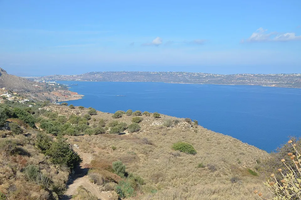 A dry hillside of scrub and a footpath falling to a deep blue bay, with a long low peninsula of land across the water.