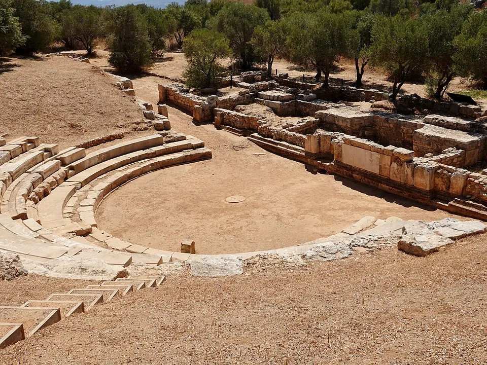 A small theatre of curved stone seat rows around a round flat orchestra, with the squared-stone walls of the stage building on the far side and olive trees behind.