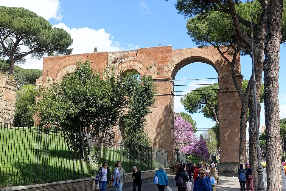 Tall red-brick arches stand behind railings and trees beside a street lined with umbrella pines, with people walking along the pavement in front.