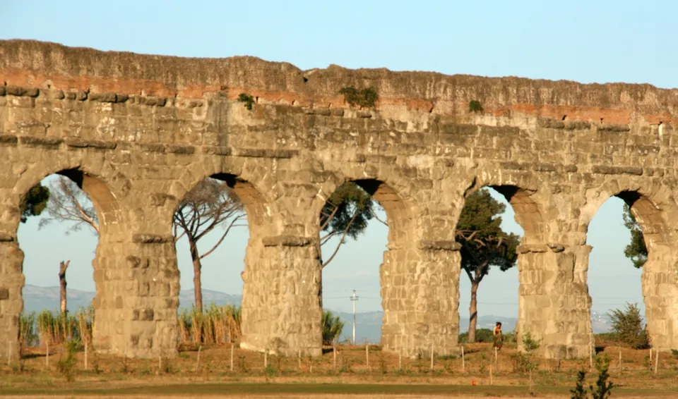 A row of tall round arches built of large weathered stone blocks, a band of reddish masonry along the top, with umbrella pines and distant hills seen through the openings.