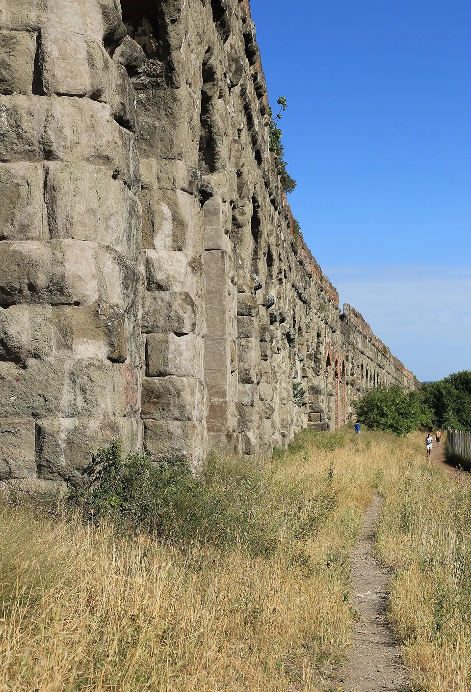 Looking along the face of the aqueduct from its foot, a wall of big squared stone blocks with plants growing in the joints, a narrow dirt path running beside it through dry grass.
