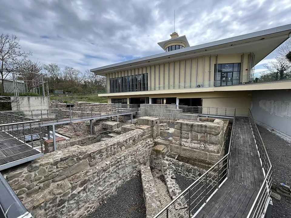 Excavated walls of rough stone laid out below raised metal walkways, with a modern two-storey museum building with a small lantern tower behind them under a cloudy sky.