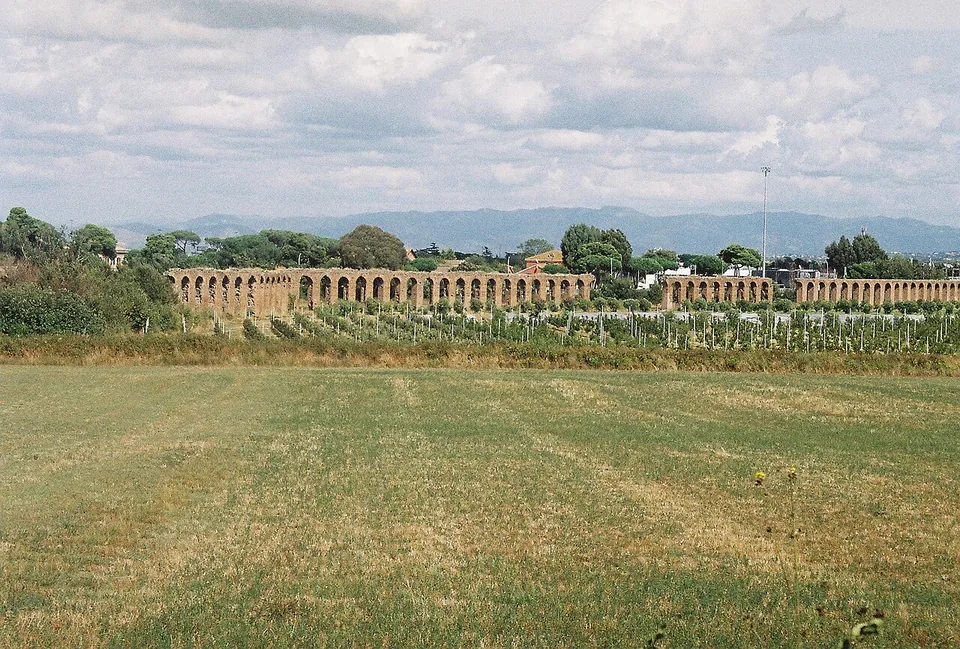 A long, level line of arches crosses the far side of a mown field, beyond a vineyard and trees, with a range of hills under cloud on the horizon.