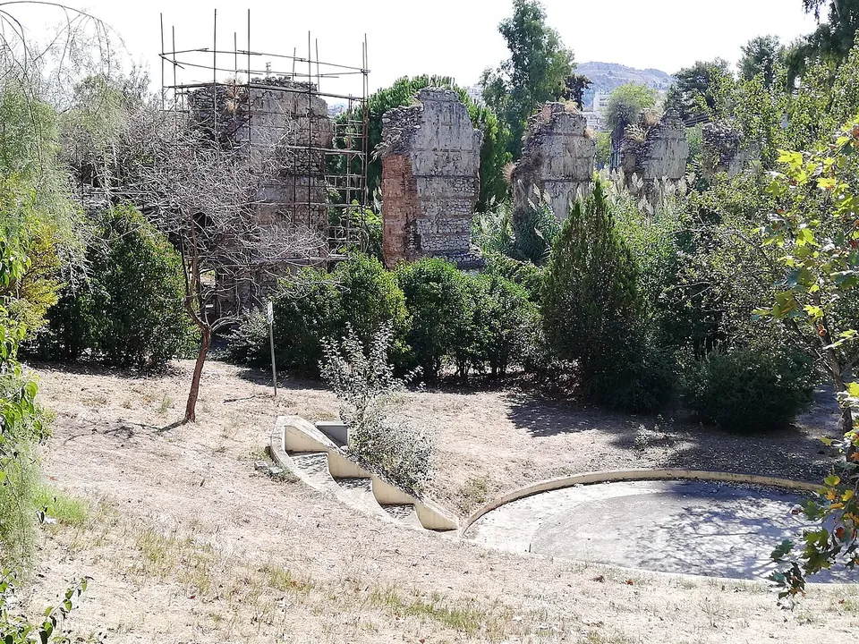 A row of ruined piers, one under scaffolding, rising behind trees and bushes above a small open-air amphitheatre in a dry park.