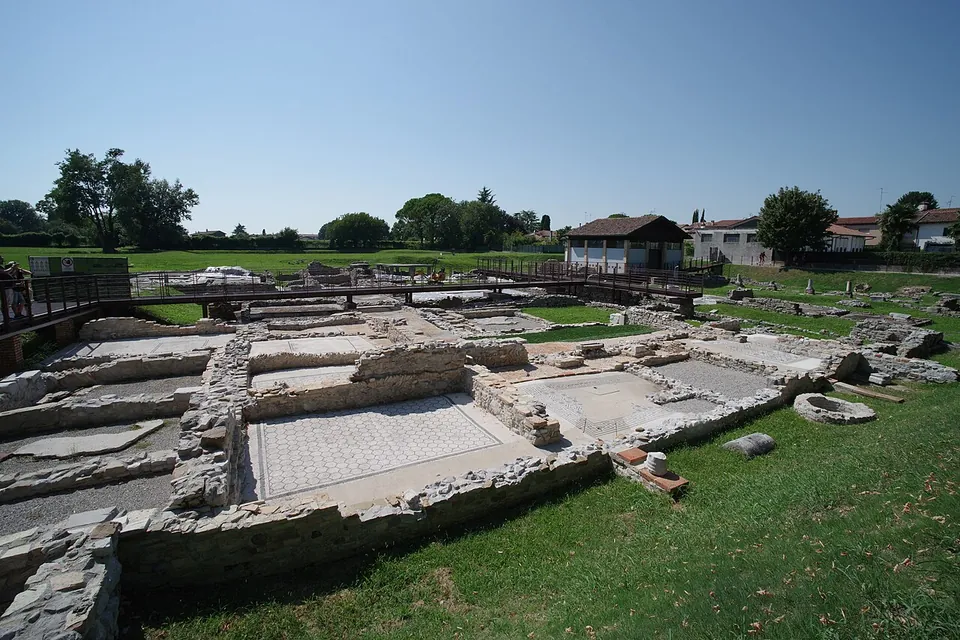 Excavated foundations of houses on a lawn, low stone walls framing rooms, one with a mosaic floor, seen from beside a wooden walkway, with trees and a small building beyond.