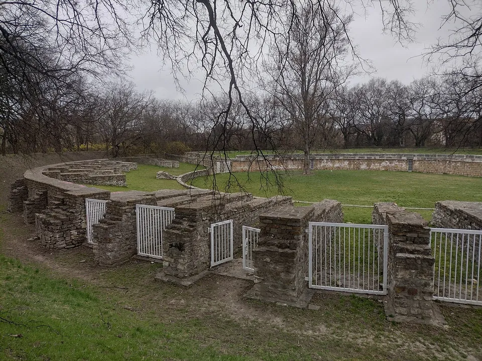 The curving outer walls of an amphitheatre in rough stone, with white metal gates closing the openings into the grassed arena, bare trees overhead.
