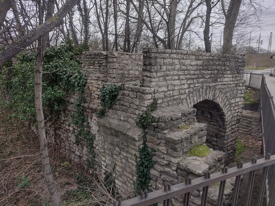 A block of rough limestone masonry with a low round-headed arch at its foot, overgrown with ivy among bare trees and behind an iron railing.