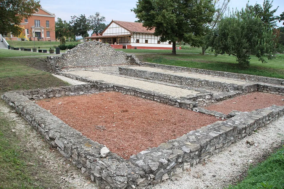 Low rough-stone walls enclosing a rectangular room floored with reddish gravel, with narrower raised strips along its sides and a heap of stones at the far end; a half-timbered house stands behind.