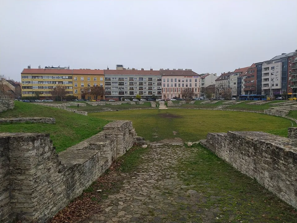 Looking down a stone-paved entrance passage between two rough limestone walls into a wide grassed arena, its far side lined with low wall stubs and, beyond them, a row of apartment buildings.