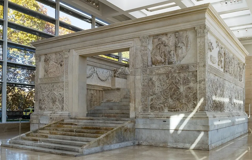 A walled marble enclosure standing in a glass-walled hall, a broad flight of steps leading up to a doorway in its front; the panels beside the doorway are carved with figures above and dense scrolling foliage below, and the altar's own steps show through the opening.
