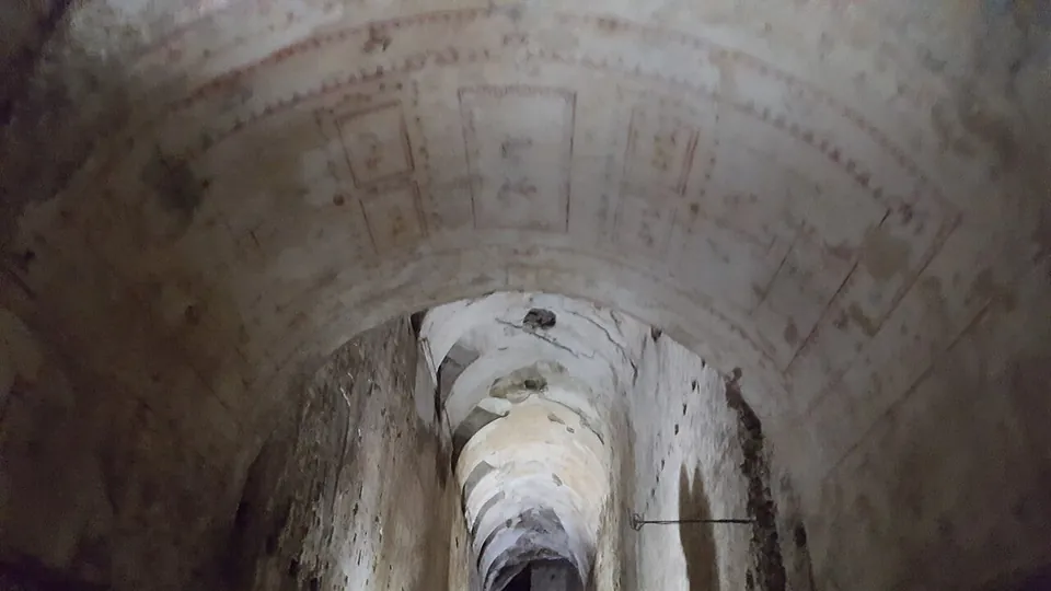 A long barrel-vaulted corridor seen from inside, faint painted panels on the vault nearest the camera and a line of lit arches receding into the distance.