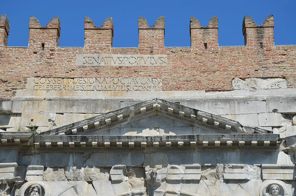Close view of the top of the arch, a carved stone pediment above a cornice, with lines of inscription in Roman capitals on the stone and brickwork above and battlements against a blue sky.