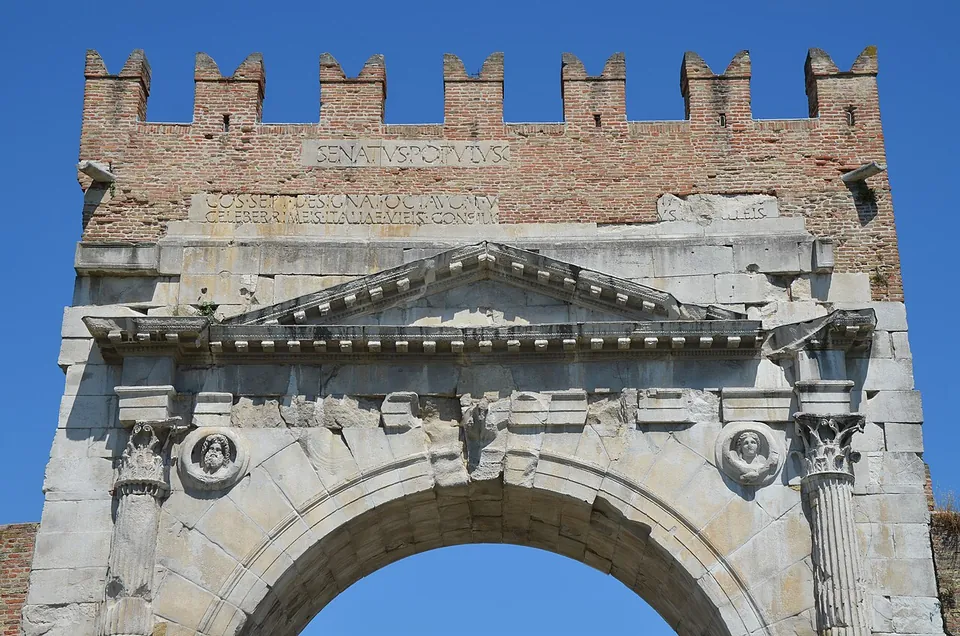 Close view of the upper arch against a blue sky - Corinthian capitals at both sides, a round medallion with a carved head in each corner above the curve of the arch, a low triangular pediment, and above it brick battlements with two lines of Roman capitals, the upper one reading SENATVS POPVLVS.