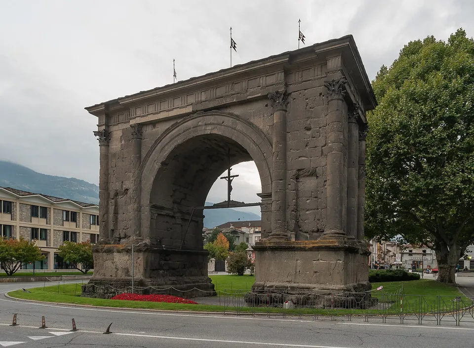 A free-standing single arch of dark grey stone on a lawn in a roundabout, its front and sides decorated with engaged columns with carved capitals, a low pitched roof on top and a crucifix hanging inside the vault, with apartment blocks and a mountain behind.