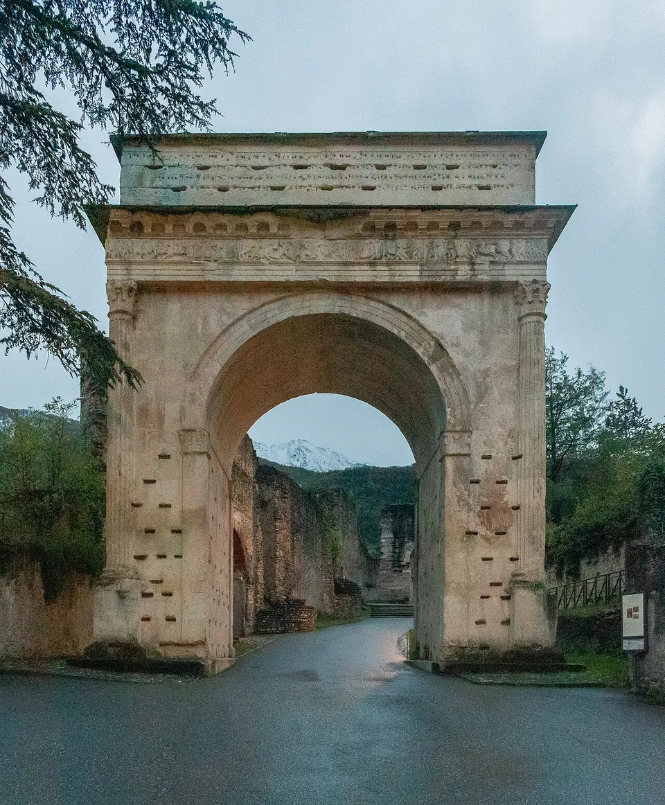 A single-arched monument of pale stone over a paved path, with fluted columns at its corners, a carved band running below the cornice and a tall plain attic on top, trees and old walls behind it.