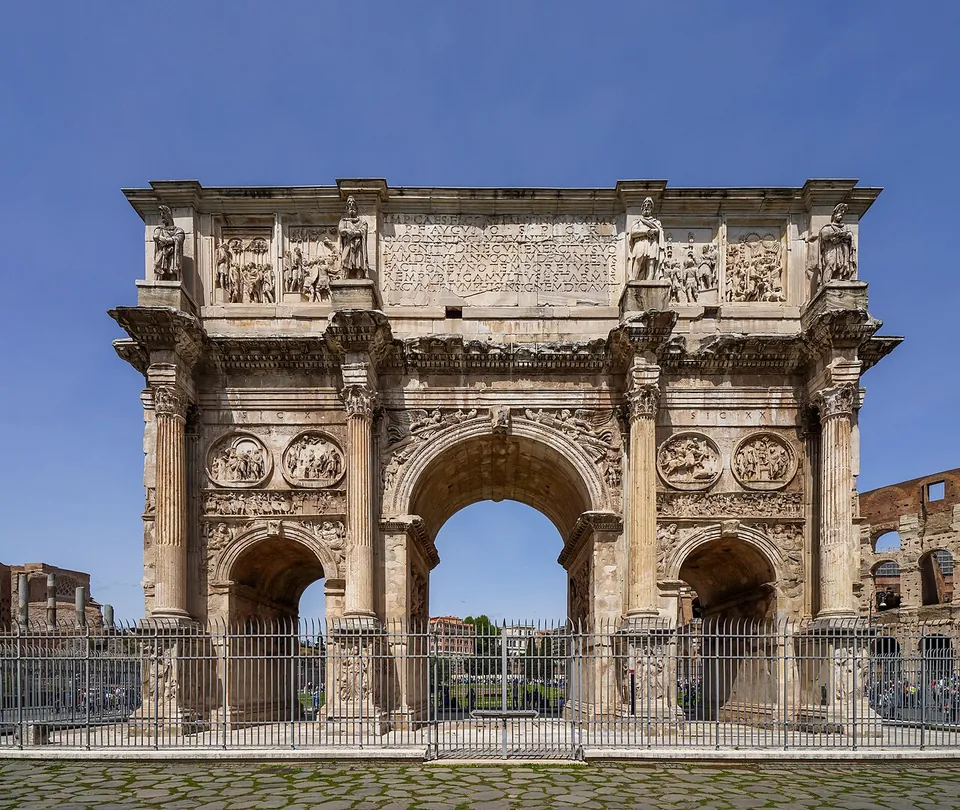 A triumphal arch with three passages seen face-on behind a modern iron railing, with detached columns, round relief panels above the side passages and statues and relief panels along the top storey; a corner of the Colosseum shows at the right edge.