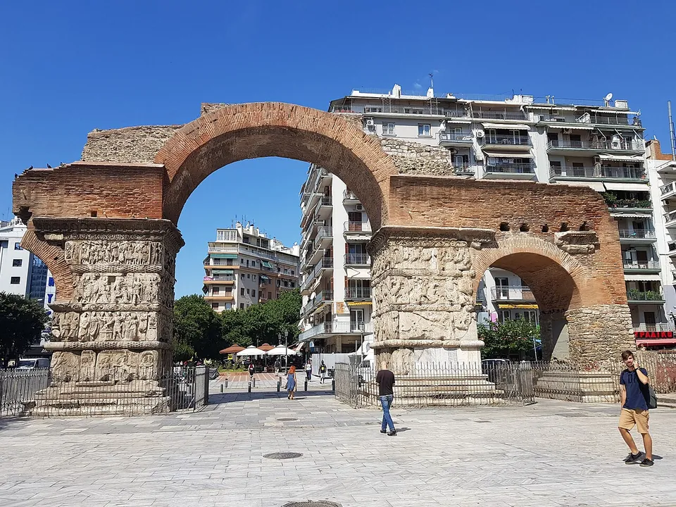 A great brick arch springing from two thick piers faced with bands of carved marble relief, with a lower brick arch to the right, standing over a wide paved pedestrian street with people walking through and apartment blocks behind.