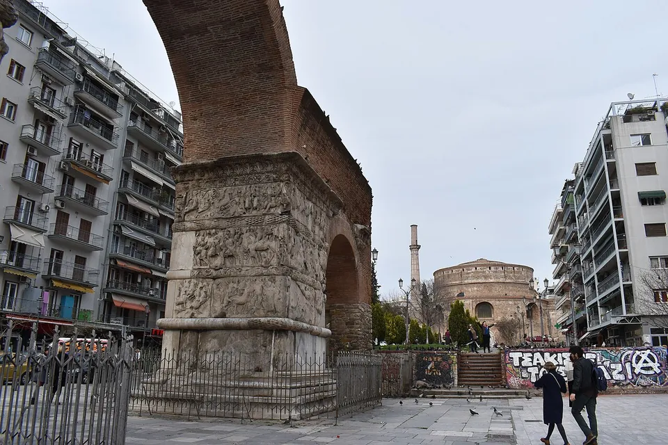 A massive brick arch on a marble pier carved with bands of relief, standing on a pedestrian street, with a domed round brick building and a minaret at the end of the street behind it.