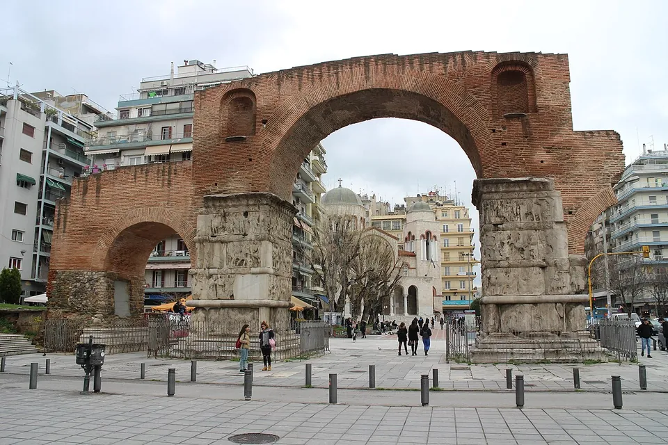 The brick arch and its relief-covered piers at the end of a paved square lined with bollards, with people walking beneath it and a domed building in the street beyond.