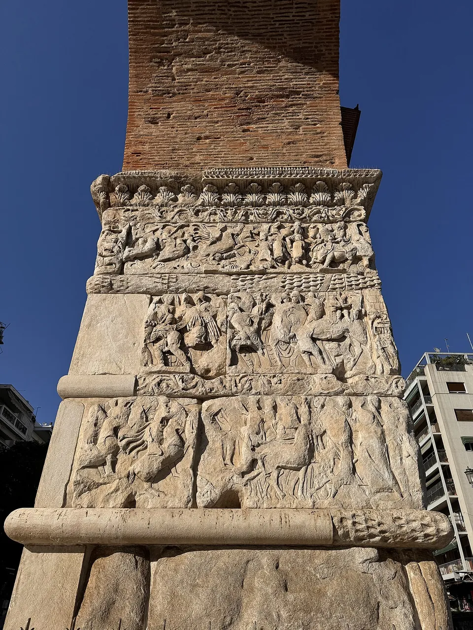 A pier seen from below, its lower part faced with marble in four carved bands of figures, horses and fighting, rising to bare brickwork above against a deep blue sky.