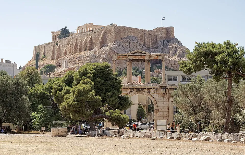A two-storey stone gateway, a round arch below and a row of columns with a small pediment above, standing among pines on open dusty ground, with the walled rock of the Acropolis and its temples behind it.