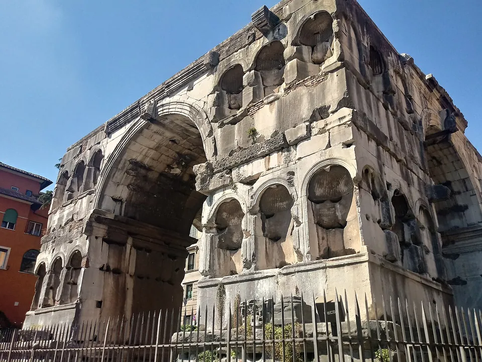 A massive four-sided arch of weathered marble blocks, its piers lined with rows of empty round-headed niches, seen from the corner behind a spiked iron railing, with a pink house beside it.