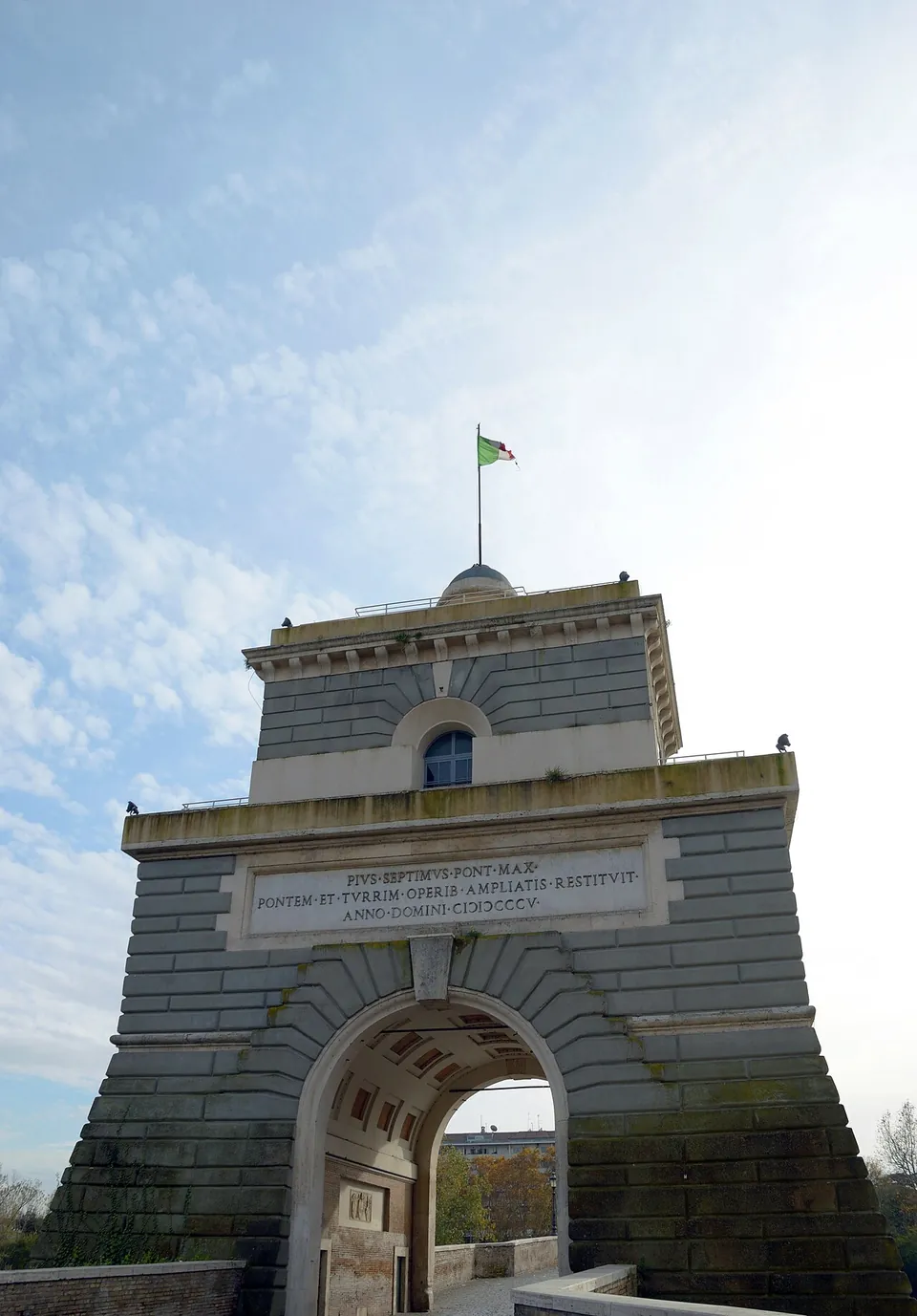 A grey stone gate tower seen from below, an arched passage through its base, a carved Latin inscription above the arch naming Pius VII and the year 1805, and a small domed top with an Italian flag.