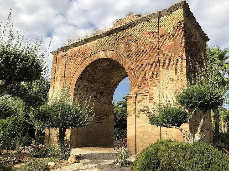 A tall single arch built of small orange-brown bricks, its piers and upper edge worn and patched, standing in a planted garden with young olive trees and palms, a gravel path running through the opening.