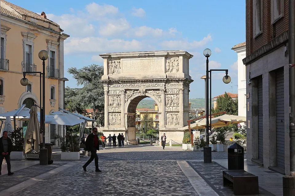 A paved pedestrian street lined with lamp posts, café umbrellas and old houses, with people walking towards a pale marble arch that closes the end of the street against trees and sky.