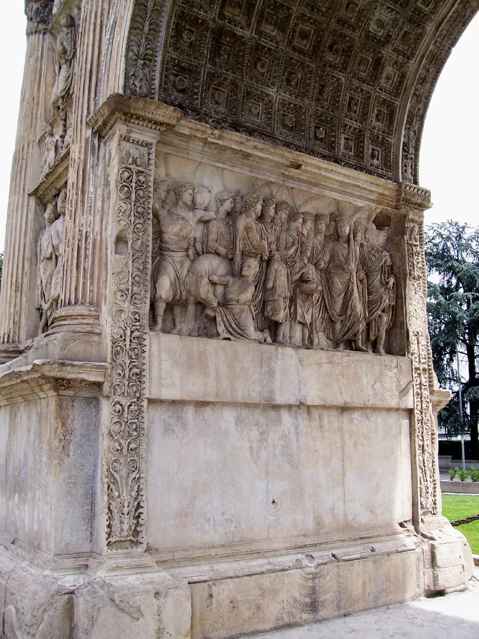 A close view of one inner face of the archway, a crowded relief of standing robed figures above a plain block of marble, framed by carved ornamental pilasters, under the coffered vault of the arch.