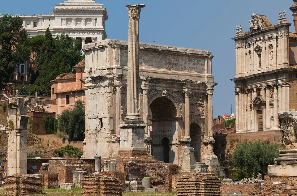 A large triple arch of weathered white marble with a lettered attic and columns on its face, a tall single column on a brick and stone base standing in front of it, and a church facade to the right.