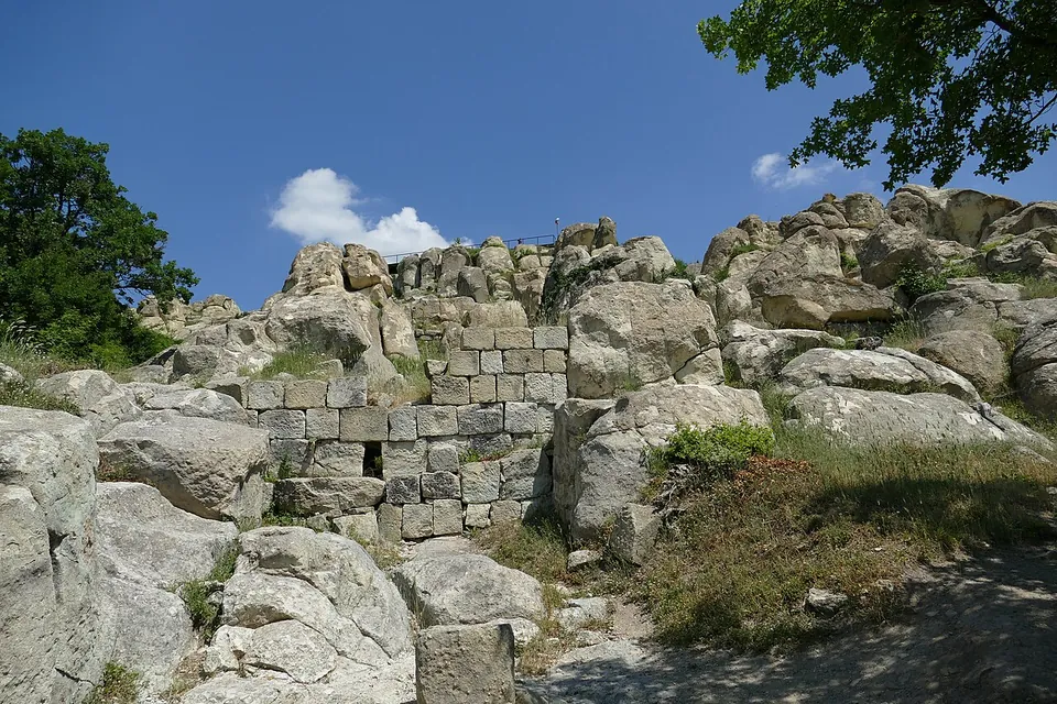 A path climbing among pale boulders towards a stretch of wall of squared blocks set into the rocks, with a tree and blue sky behind.