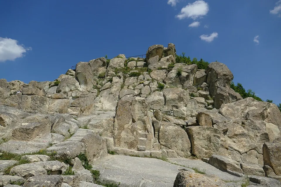 A great outcrop of pale grey rock rising to a summit, its surface cut with steps, ledges and flat platforms, under a blue sky with a few clouds.