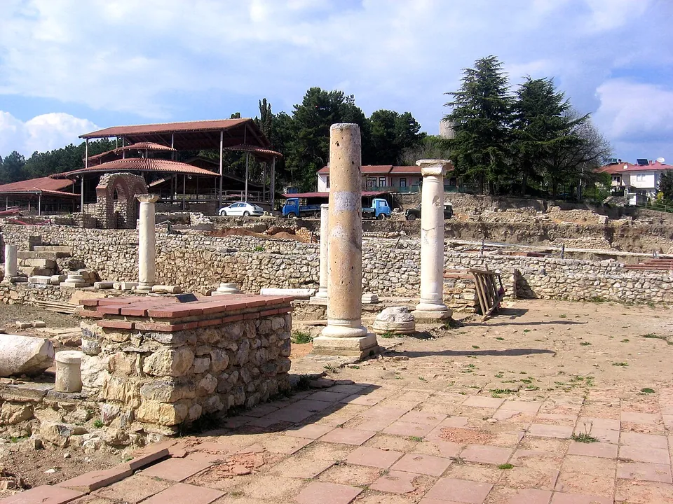 Excavated low stone walls, two columns standing on their bases and a paved brick floor in bright sun, with roofed shelters and trees behind.
