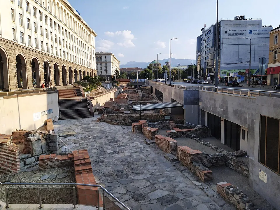 A sunken archaeological area paved with large irregular stones and crossed by low brick and stone walls, below street level, with steps leading down from a colonnaded government building on the left and traffic on a boulevard on the right.